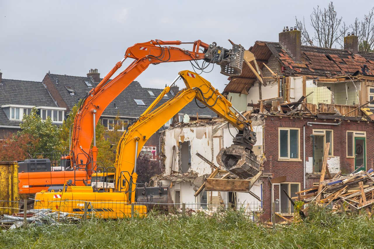Twee sloopkranen halen een rij bakstenen woningen neer, met de kap en de verdiepingsvloeren al open.